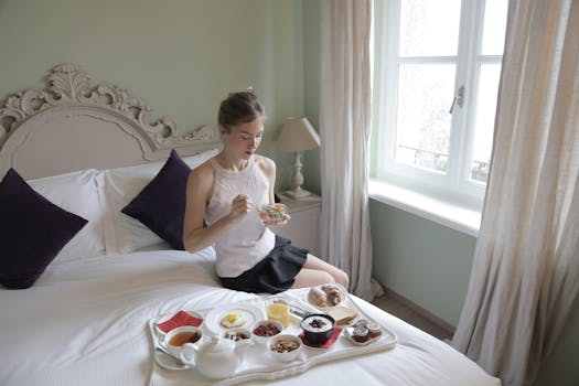 Femme dégustant un petit-déjeuner sur un lit dans une chambre lumineuse avec des fenêtres et des rideaux clairs.