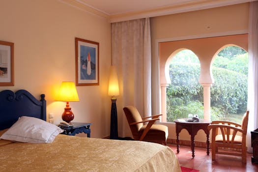 Chambre d'hôtel avec lit, fauteuils, table et fenêtre donnant sur un jardin verdoyant. Ambiance chaleureuse et accueillante.